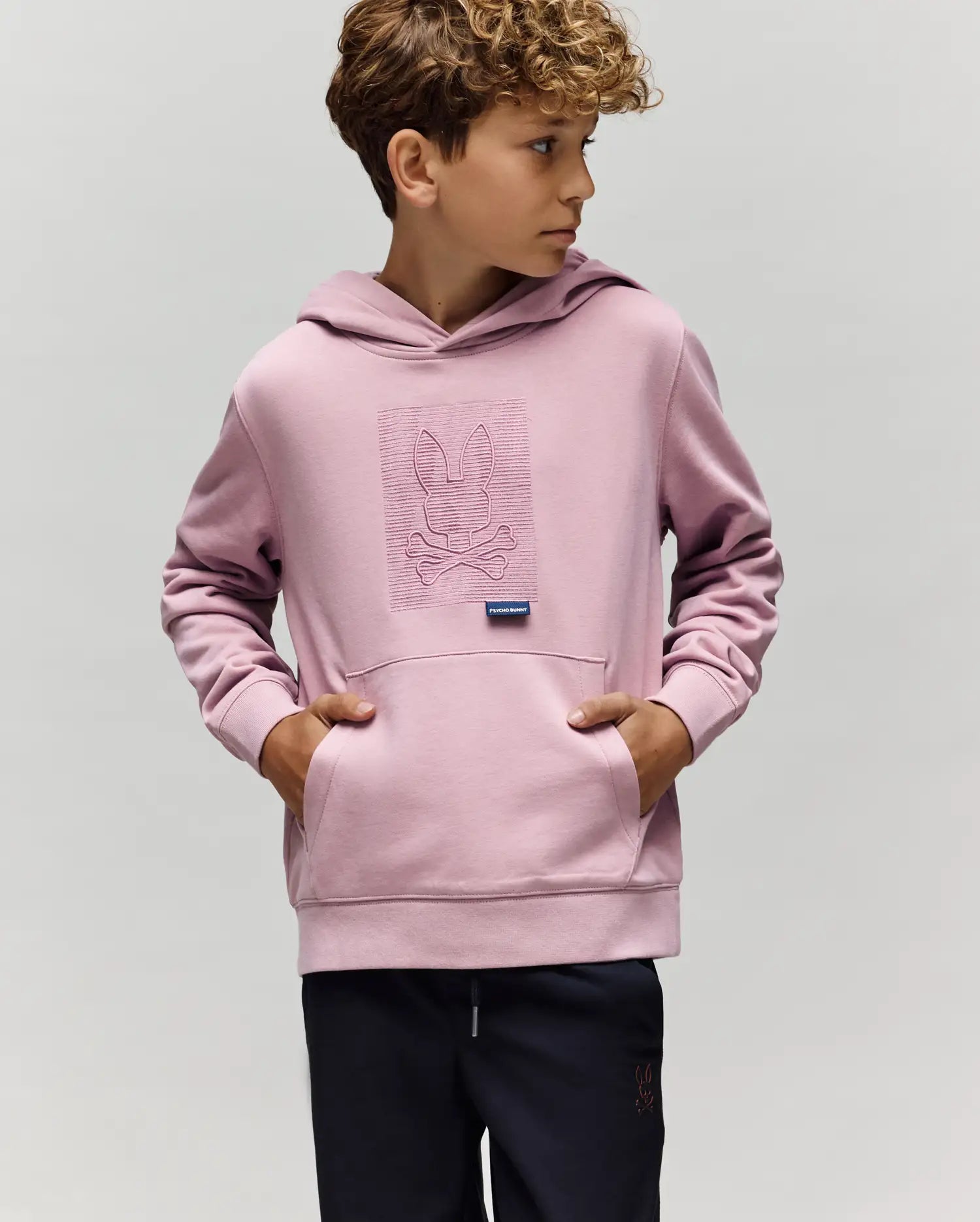 A young boy wears the KIDS GRANT HOODIE - KT0500020 by Psycho Bunny in light purple, made from a premium cotton blend with an embroidered Bunny skull and crossbones, standing with hands in the pocket against a plain background.