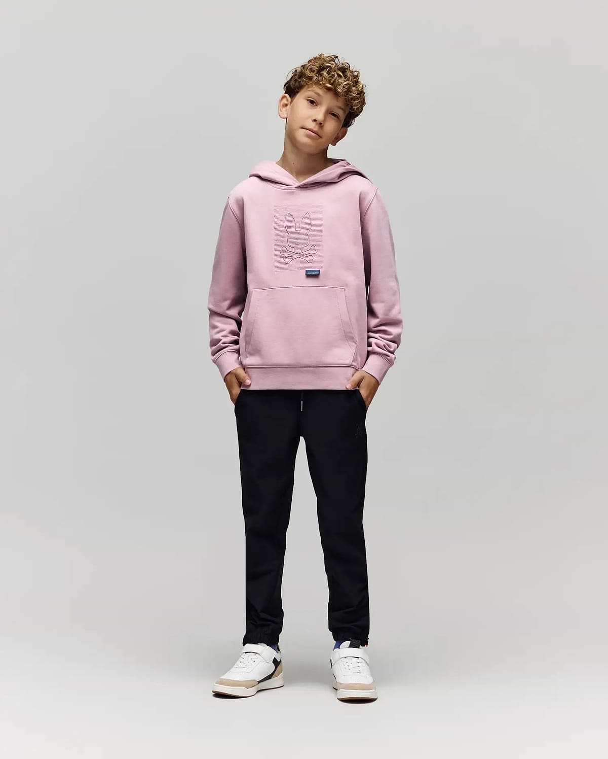 A young boy with curly hair stands against a plain background, hands in pockets, wearing the Psycho Bunny KIDS GRANT HOODIE (KT0500020), black pants, and white sneakers while looking at the camera.