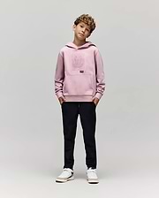 A young boy with curly hair stands against a plain background, hands in pockets, wearing the Psycho Bunny KIDS GRANT HOODIE (KT0500020), black pants, and white sneakers while looking at the camera.