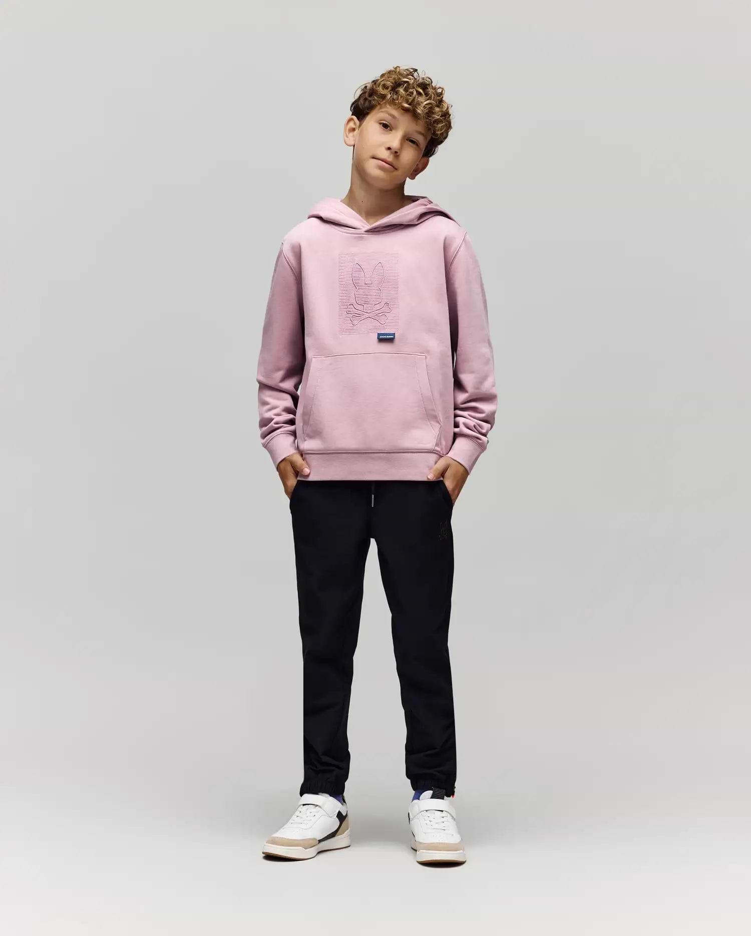 A young boy with curly hair stands against a plain background, hands in pockets, wearing the Psycho Bunny KIDS GRANT HOODIE (KT0500020), black pants, and white sneakers while looking at the camera.