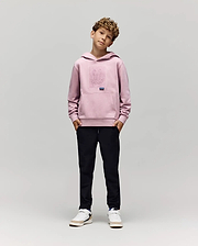 A young boy with curly hair stands against a plain background, hands in pockets, wearing the Psycho Bunny KIDS GRANT HOODIE (KT0500020), black pants, and white sneakers while looking at the camera.