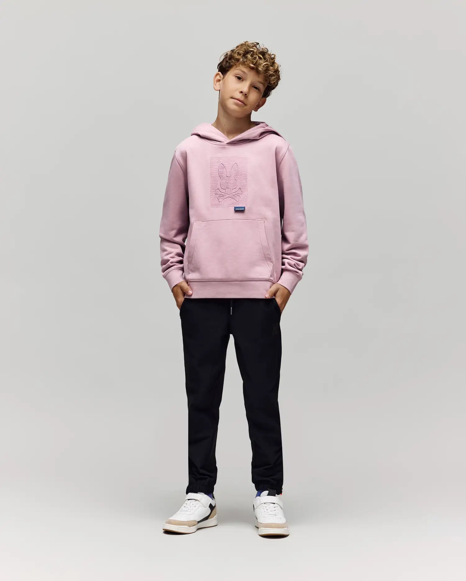 A young boy with curly hair stands against a plain background, hands in pockets, wearing the Psycho Bunny KIDS GRANT HOODIE (KT0500020), black pants, and white sneakers while looking at the camera.