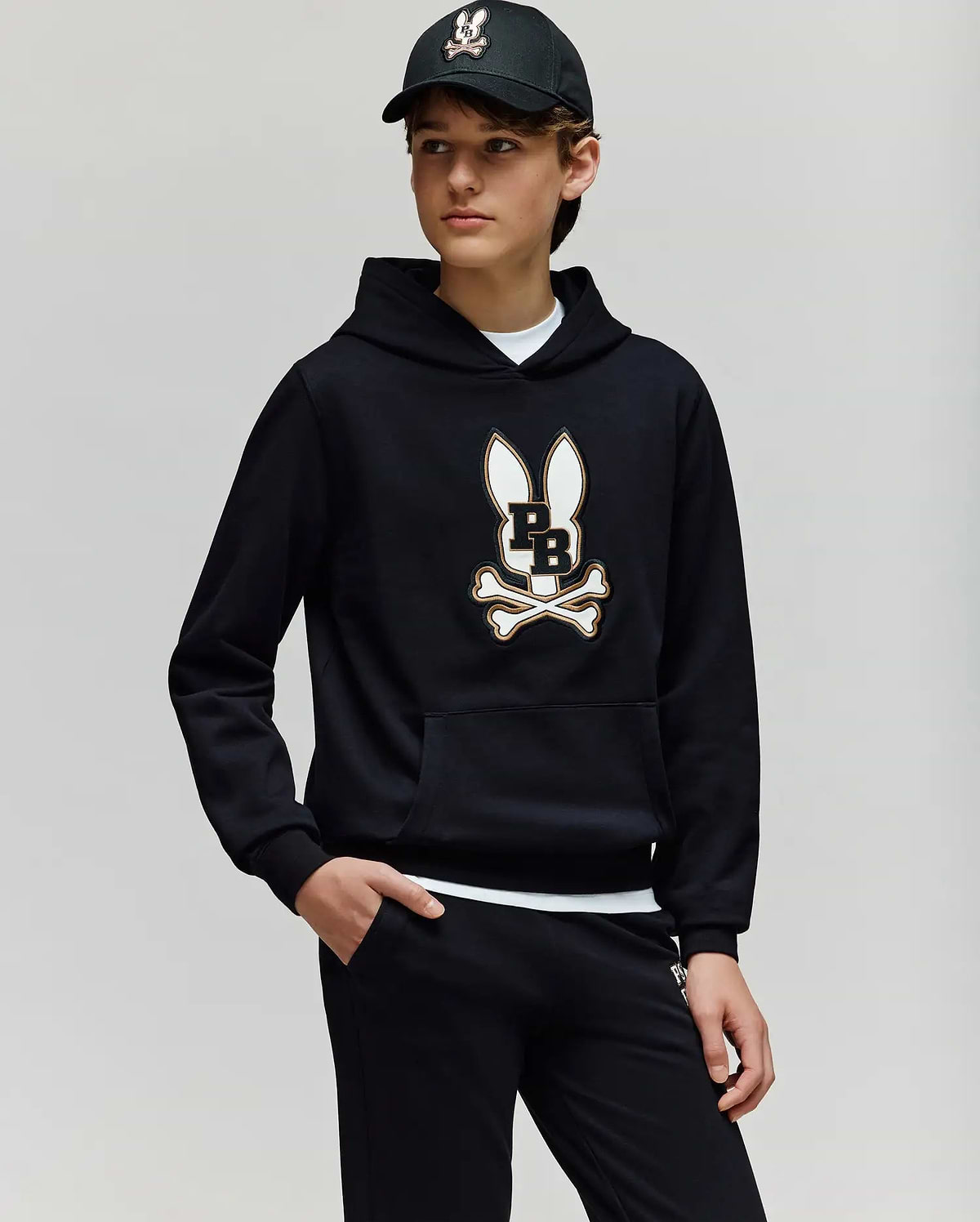 A teen boy wears the Psycho Bunny KIDS WADE CORD PATCH HOODIE (KT0500031), showcasing its premium cotton-blend fabric and signature Bunny patch with crossbones, paired with black pants and a black baseball cap.