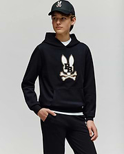 A teen boy wears the Psycho Bunny KIDS WADE CORD PATCH HOODIE (KT0500031), showcasing its premium cotton-blend fabric and signature Bunny patch with crossbones, paired with black pants and a black baseball cap.