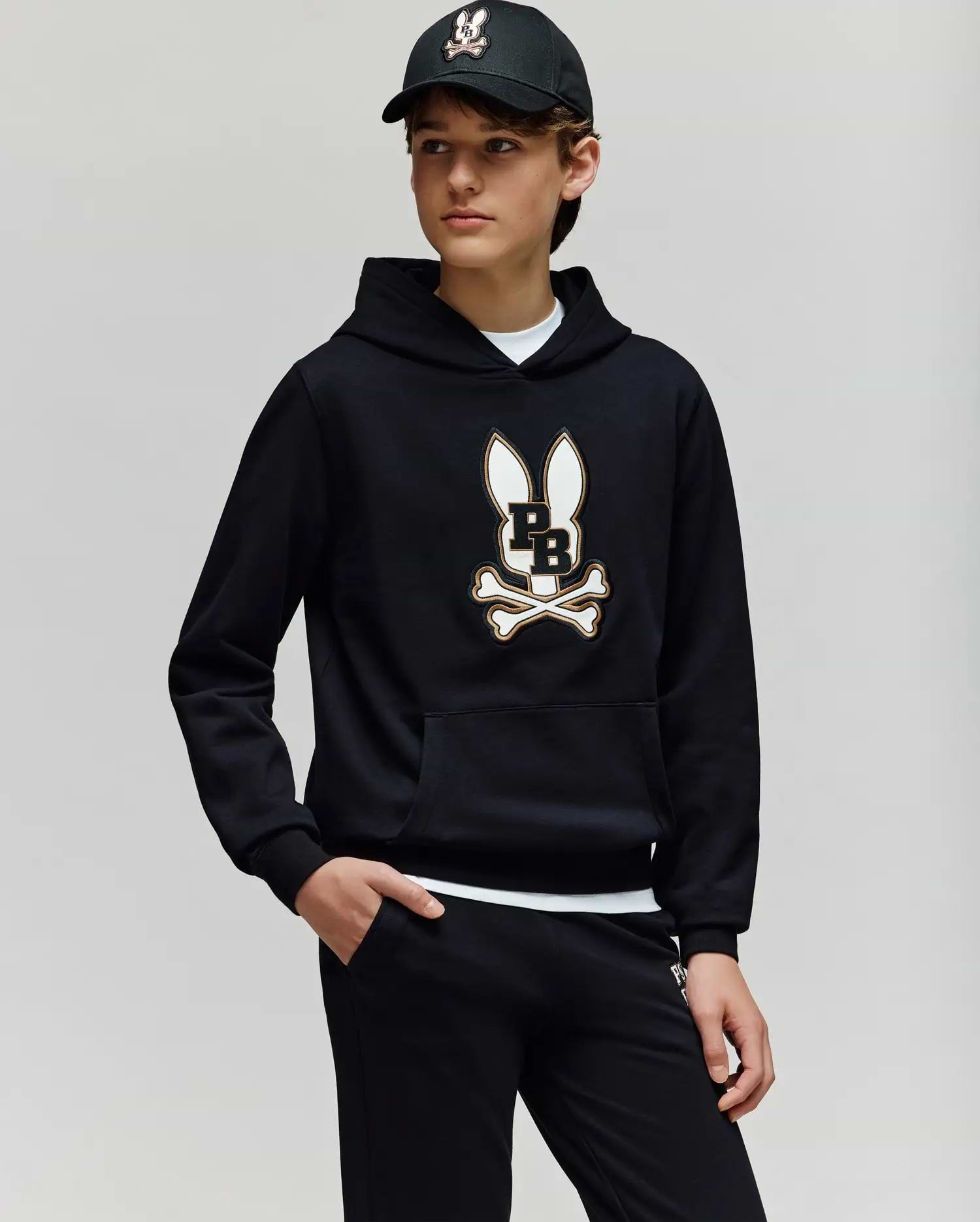 A teen boy wears the Psycho Bunny KIDS WADE CORD PATCH HOODIE (KT0500031), showcasing its premium cotton-blend fabric and signature Bunny patch with crossbones, paired with black pants and a black baseball cap.