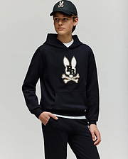 A teen boy wears the Psycho Bunny KIDS WADE CORD PATCH HOODIE (KT0500031), showcasing its premium cotton-blend fabric and signature Bunny patch with crossbones, paired with black pants and a black baseball cap.
