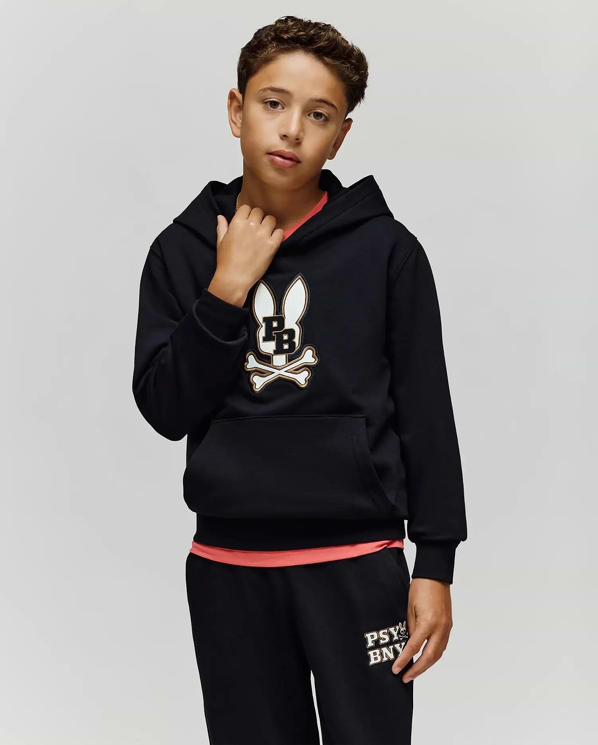 A boy wears the Psycho Bunny KIDS WADE CORD PATCH HOODIE (KT0500031) with an embroidered Bunny patch and matching black pants, standing against a plain light gray background.