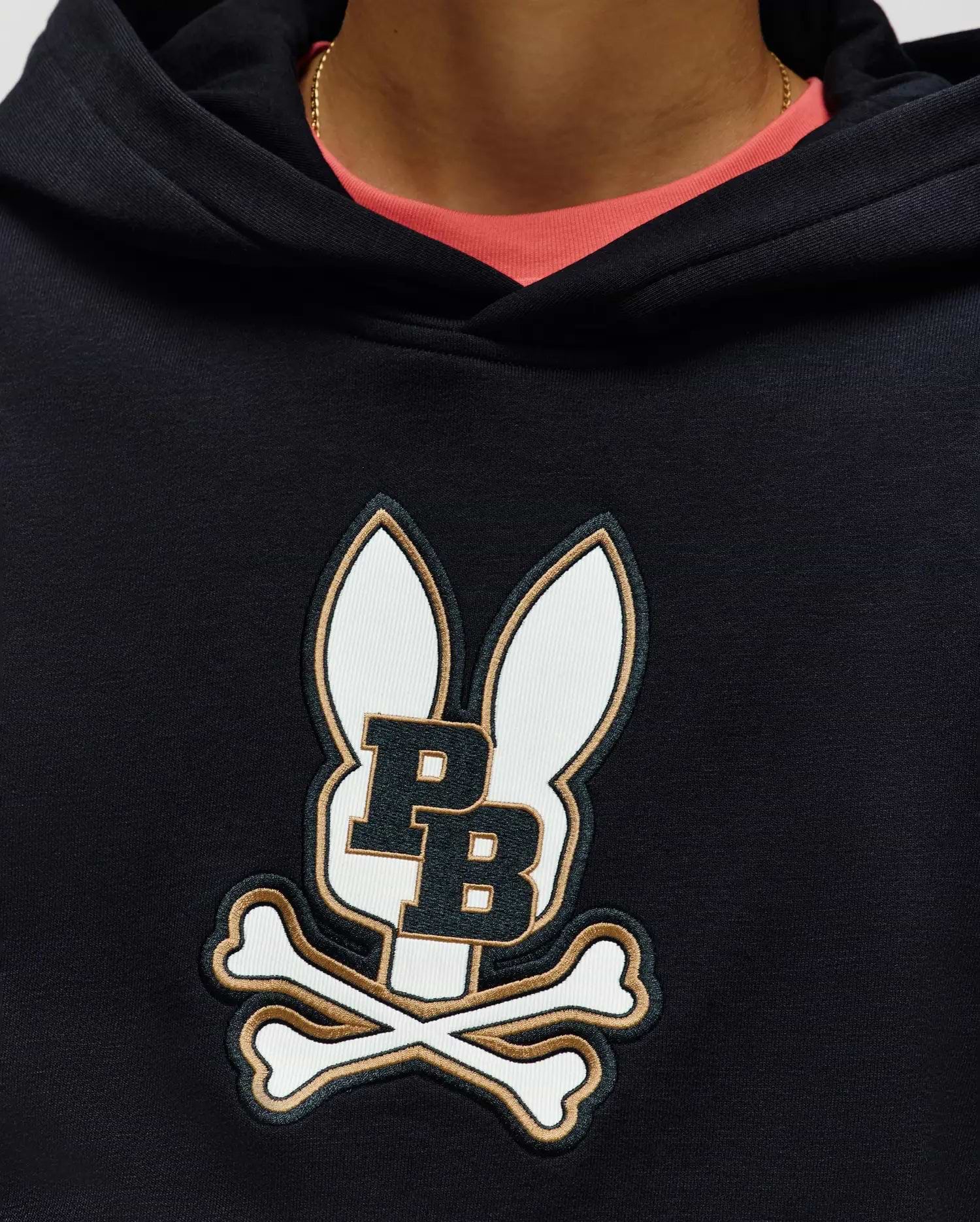 A person wears the Psycho Bunny KIDS WADE CORD PATCH HOODIE (KT0500031), made from a premium cotton blend with an embroidered Bunny patch showing bunny ears, "PB," and crossed bones. A pink shirt and gold chain peek at the neckline.