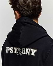 A child with short brown hair is seen from behind, wearing the Psycho Bunny KIDS WADE CORD PATCH HOODIE (KT0500031), a premium cotton blend black hoodie featuring "PSYBONY" and a small skull and crossbones in white on the back.