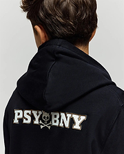 A child with short brown hair is seen from behind, wearing the Psycho Bunny KIDS WADE CORD PATCH HOODIE (KT0500031), a premium cotton blend black hoodie featuring "PSYBONY" and a small skull and crossbones in white on the back.