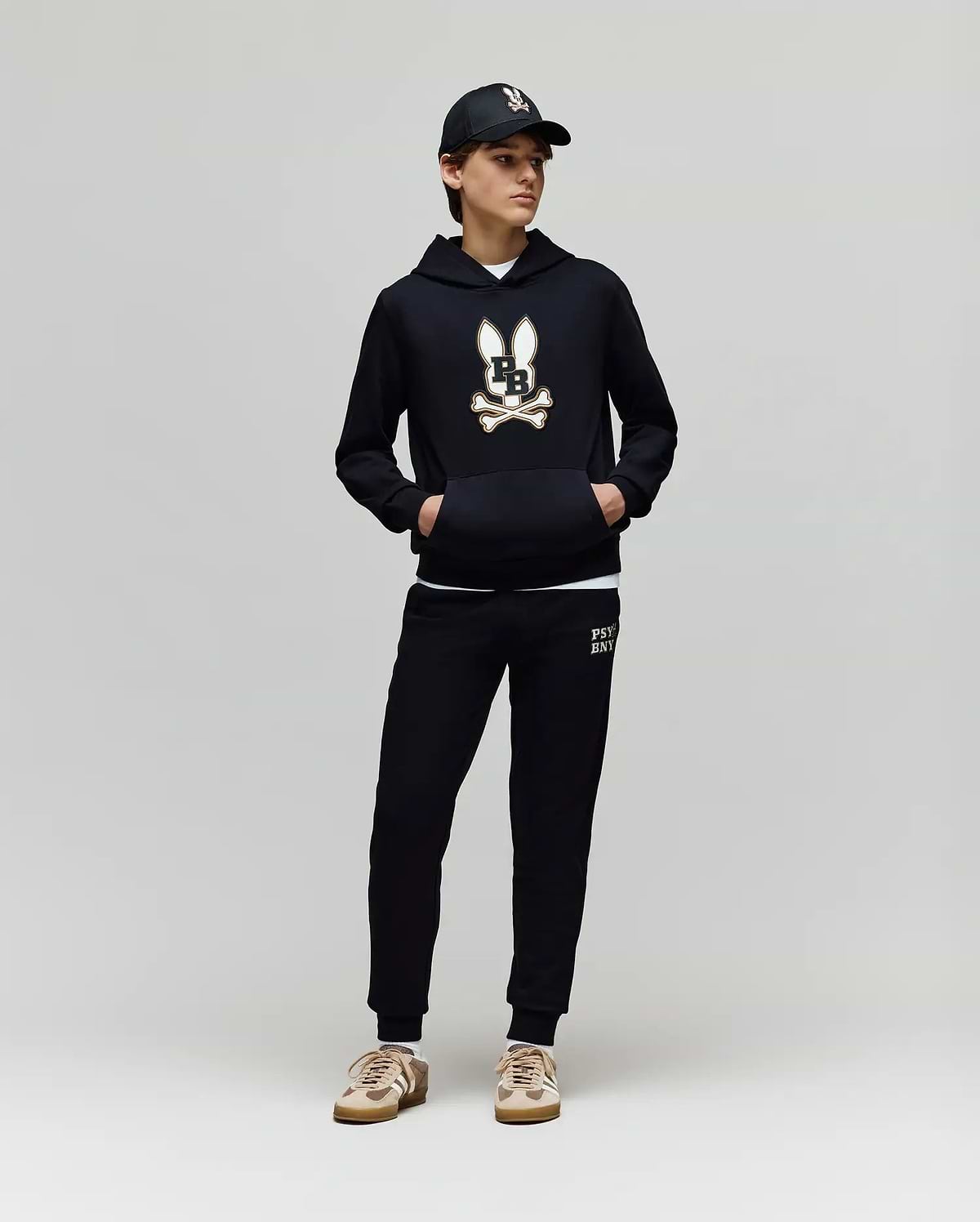 A person in a Psycho Bunny KIDS WADE CORD PATCH HOODIE (KT0500031), black joggers, black cap, and beige sneakers stands against a plain light gray background.