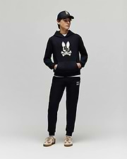 A person in a Psycho Bunny KIDS WADE CORD PATCH HOODIE (KT0500031), black joggers, black cap, and beige sneakers stands against a plain light gray background.