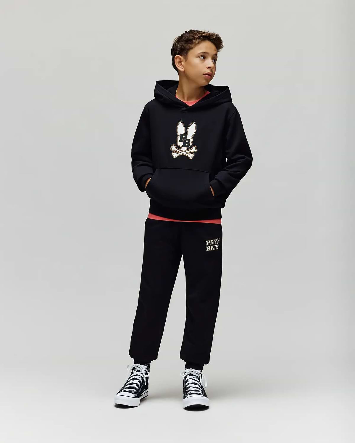 A boy stands against a plain background wearing the Psycho Bunny KIDS WADE CORD PATCH HOODIE (KT0500031), paired with black sweatpants featuring leg text and black high-top sneakers. He has his hands in his pockets and looks to the side.