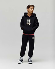 A boy stands against a plain background wearing the Psycho Bunny KIDS WADE CORD PATCH HOODIE (KT0500031), paired with black sweatpants featuring leg text and black high-top sneakers. He has his hands in his pockets and looks to the side.