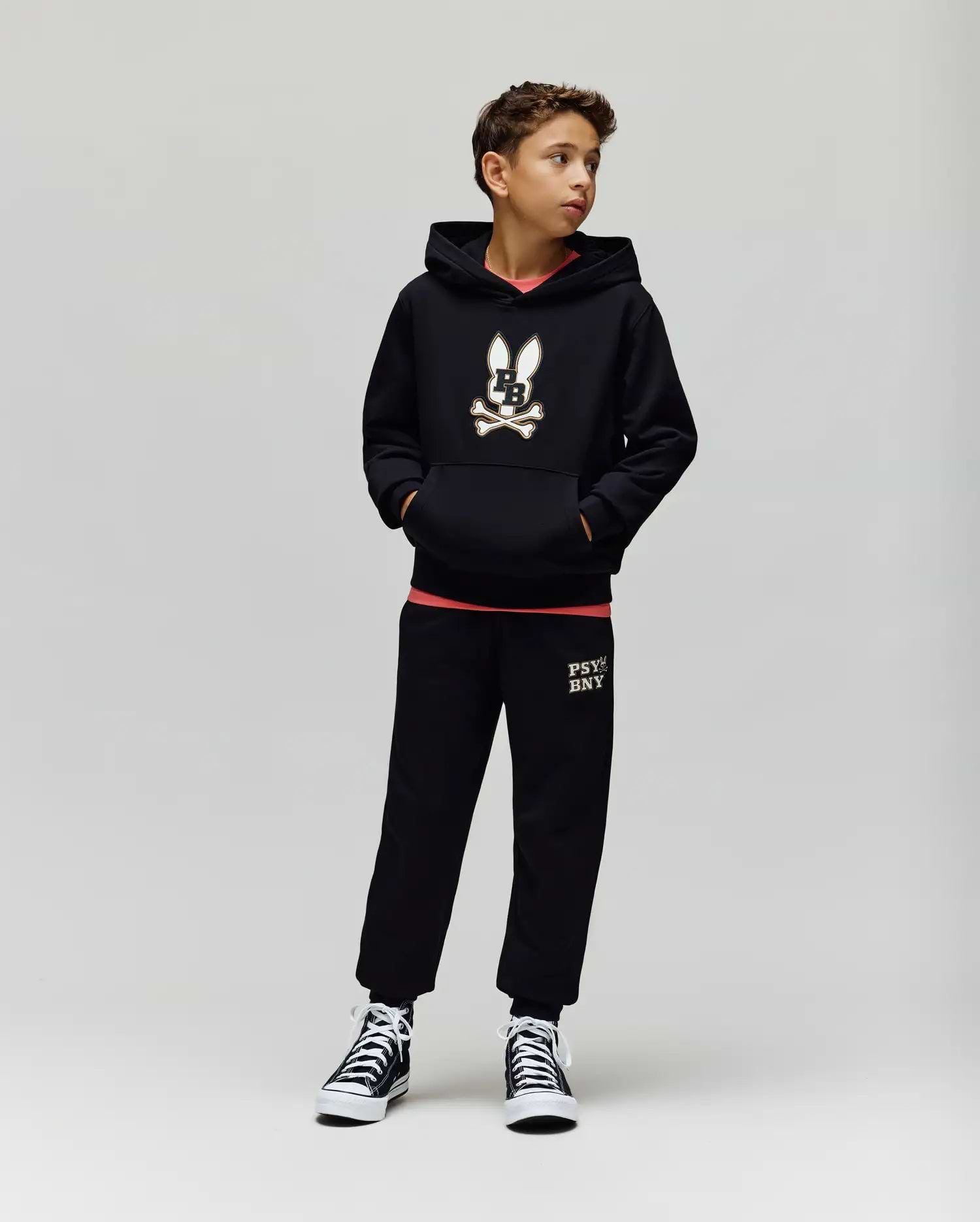A boy stands against a plain background wearing the Psycho Bunny KIDS WADE CORD PATCH HOODIE (KT0500031), paired with black sweatpants featuring leg text and black high-top sneakers. He has his hands in his pockets and looks to the side.