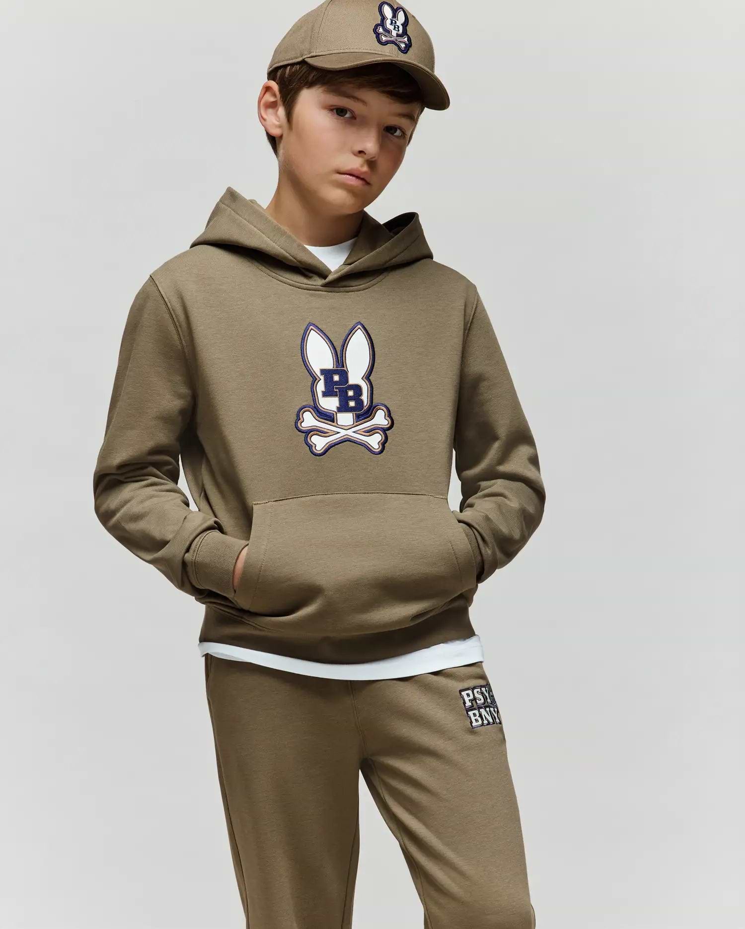 A boy wears the Psycho Bunny KIDS WADE CORD PATCH HOODIE (KT0500031), a tan premium cotton-blend hoodie with a cartoon bunny and crossbones logo, standing with hands in pockets against a plain light background.