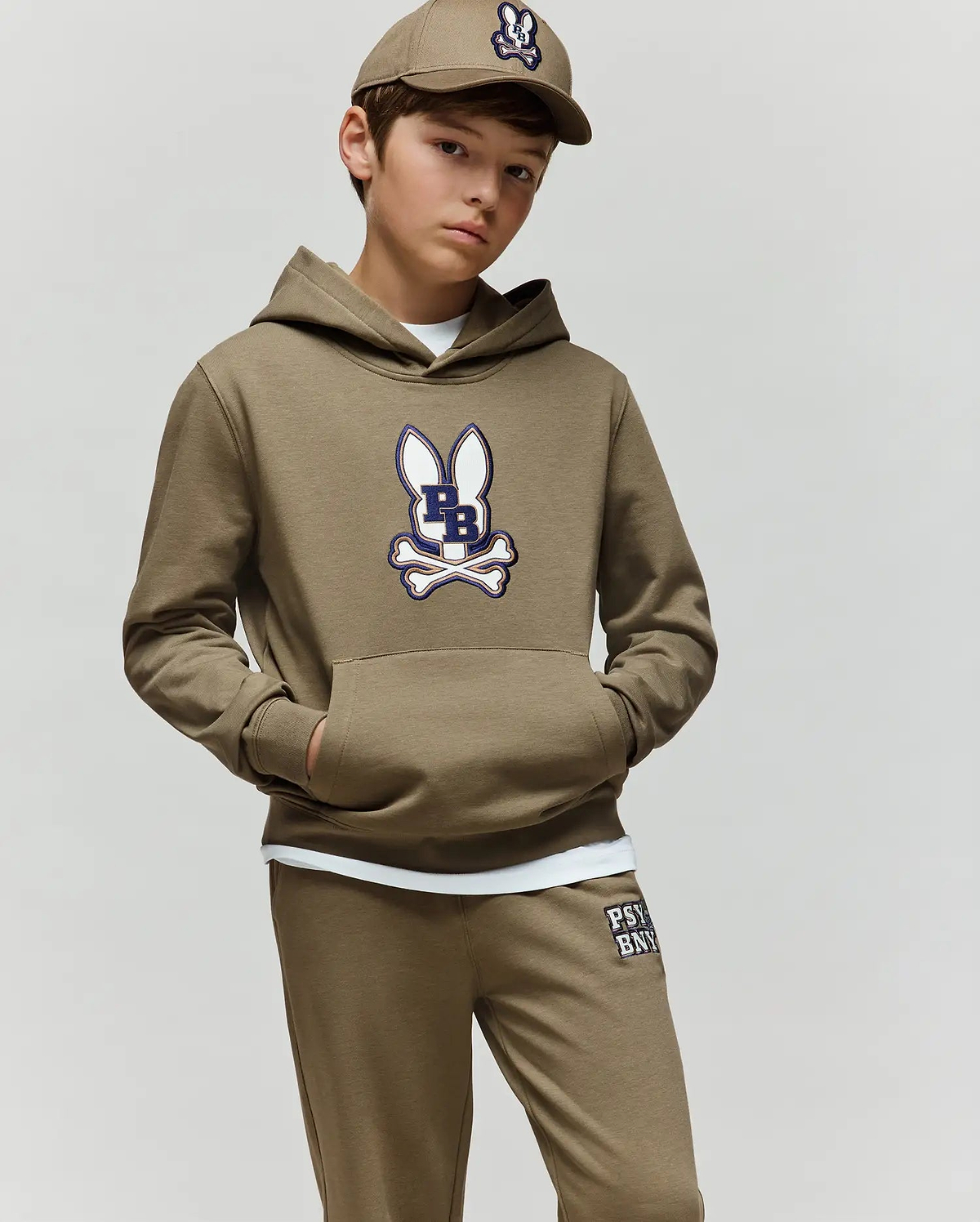 A boy wears the Psycho Bunny KIDS WADE CORD PATCH HOODIE (KT0500031), a tan premium cotton-blend hoodie with a cartoon bunny and crossbones logo, standing with hands in pockets against a plain light background.