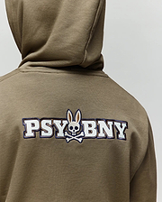 A child wears the Psycho Bunny KIDS WADE CORD PATCH HOODIE (KT0500031), an olive green pullover featuring "PSYBNY" and a skull-and-crossbones bunny logo embroidered on the back, crafted from a premium cotton blend.