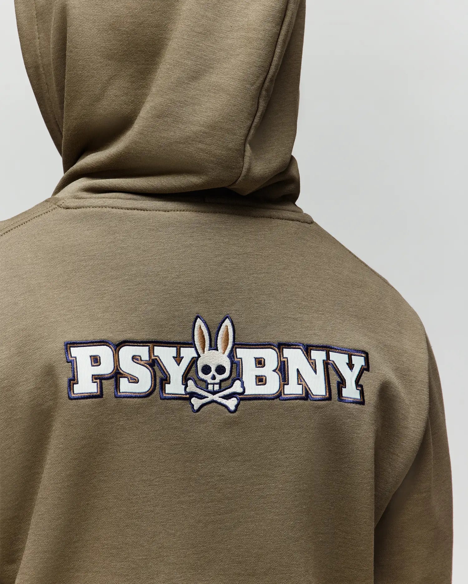 A child wears the Psycho Bunny KIDS WADE CORD PATCH HOODIE (KT0500031), an olive green pullover featuring "PSYBNY" and a skull-and-crossbones bunny logo embroidered on the back, crafted from a premium cotton blend.