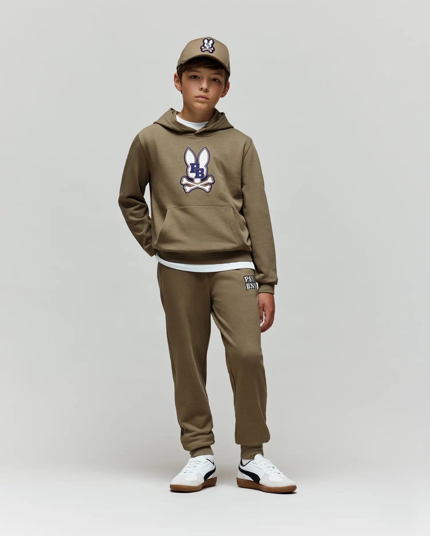 A boy in a Psycho Bunny KIDS WADE CORD PATCH HOODIE (KT0500031), olive green with cartoon bunny logo, matching sweatpants and cap, plus white sneakers, stands against a light background; crafted from premium cotton blend for comfort.