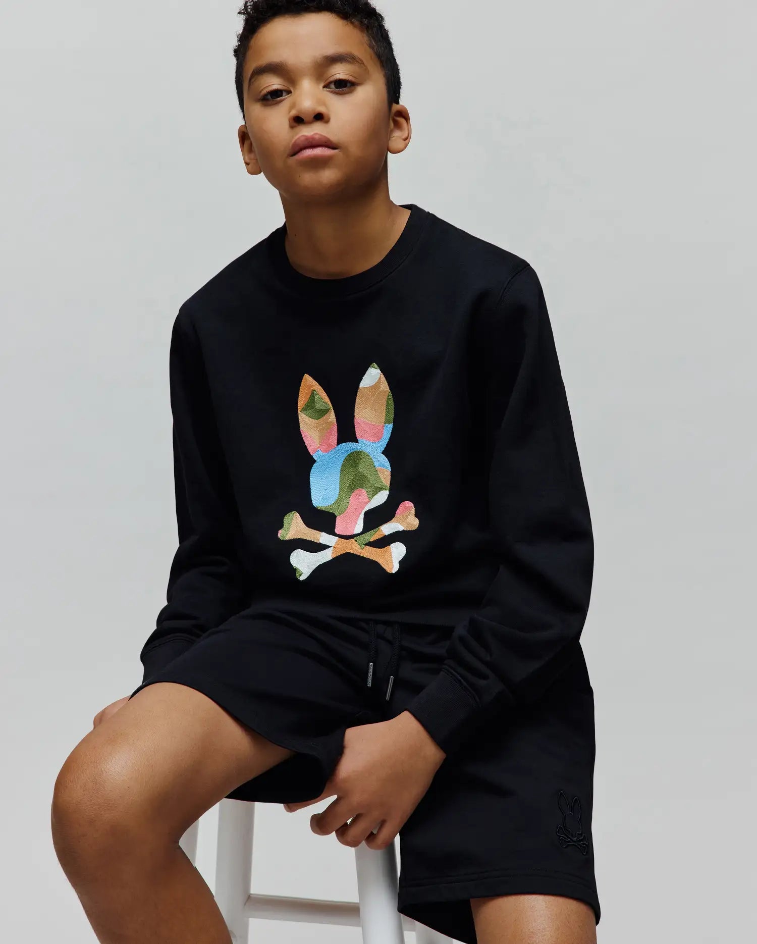 Black Kids Ara Crew sweatshirt with a large multicolor camo embroidered Psycho Bunny skull on the chest, paired with black shorts featuring a small tonal embroidered bunny skull on the left leg.