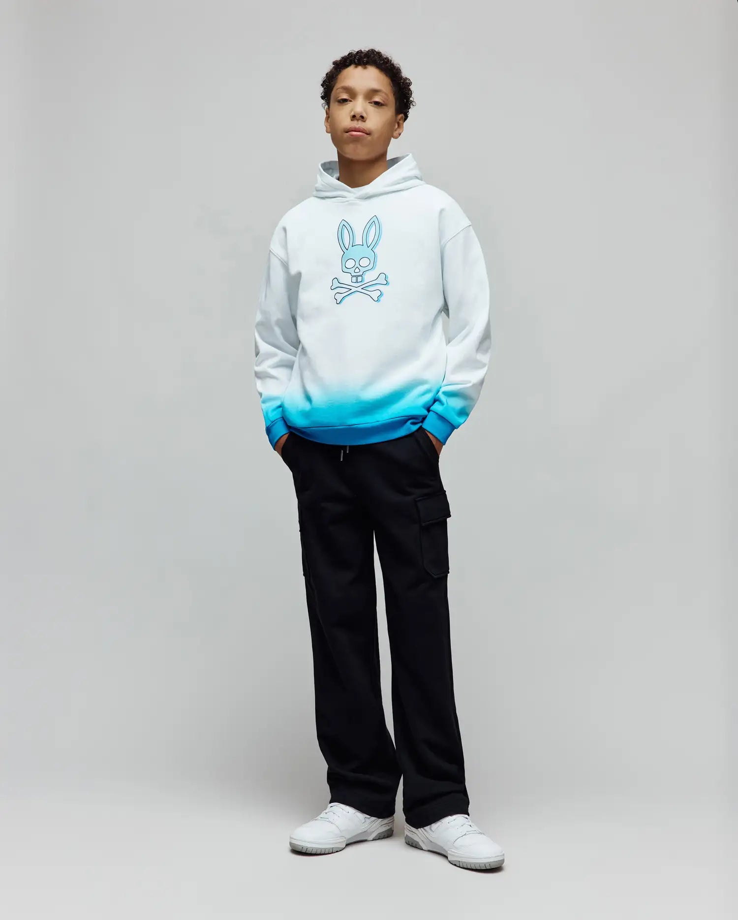 Kids Dax Oversized Hoodie in white French terry with a blue ombre fade at the hem and cuffs, featuring a large blue Psycho Bunny skull and crossbones graphic centered on the chest.