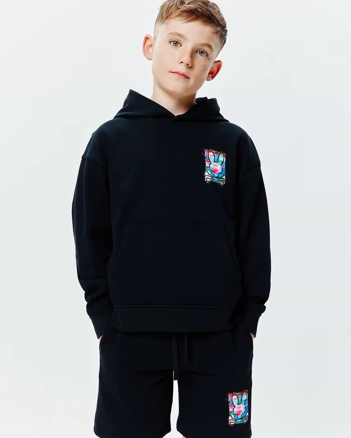 Black Kids Isaac Print Hoodie and matching shorts, relaxed fit, soft jersey. Both feature a small, vibrant, multi-color abstract Psycho Bunny skull logo on left chest and left thigh.