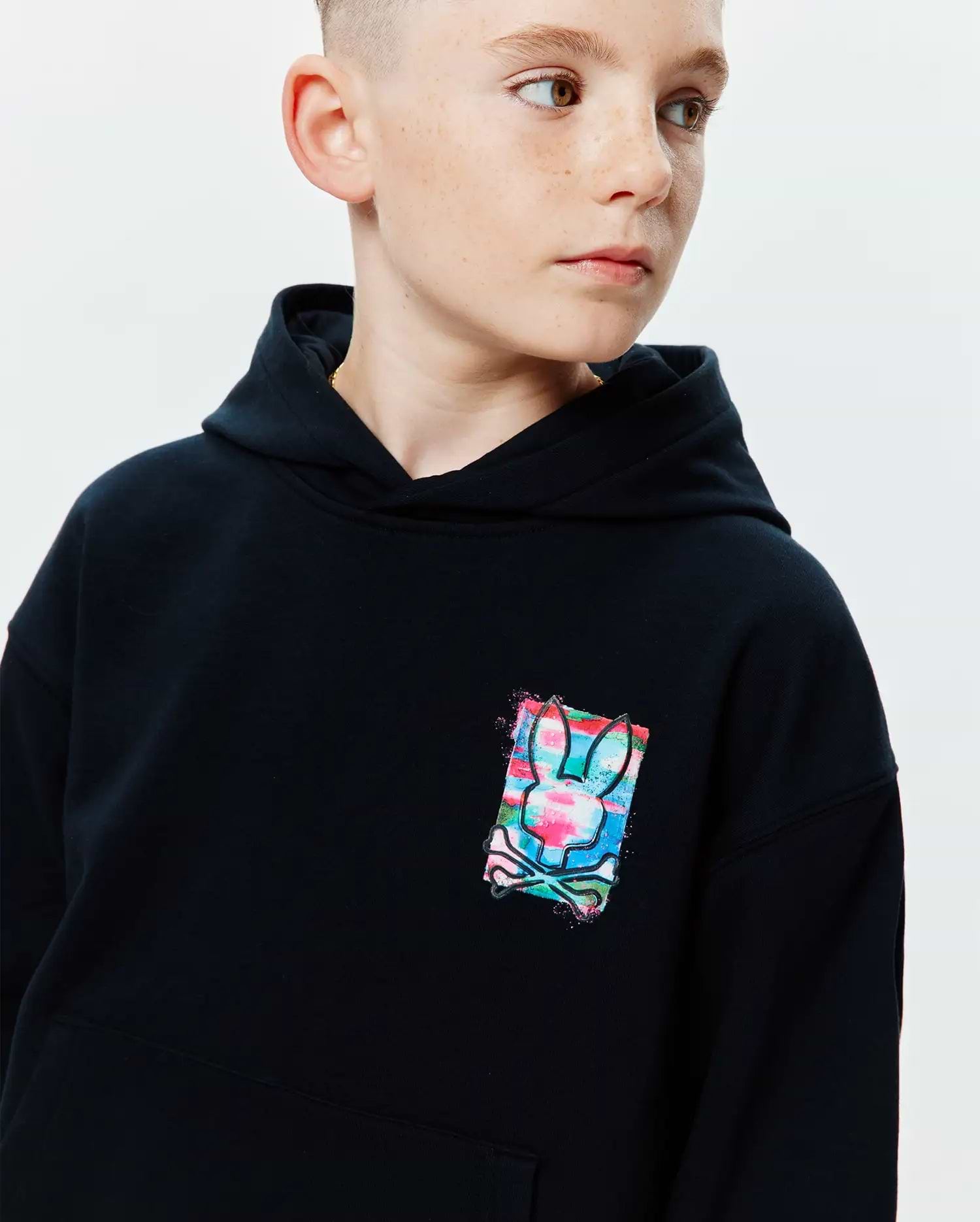 Kids Isaac Print Hoodie, black relaxed fit jersey hoodie with a small, vibrant multi-color graphic print of the Psycho Bunny skull logo on the chest, featuring pink, blue, and green splashes.