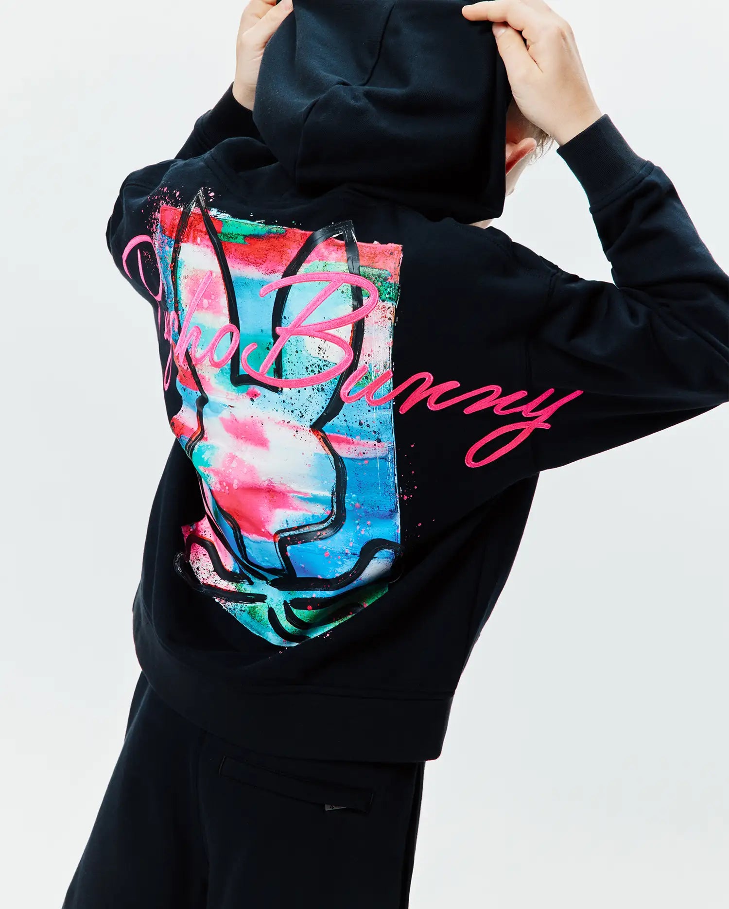 Kids Isaac Print Hoodie, black relaxed fit French terry with a large, vibrant paint-splatter Psycho Bunny skull graphic and pink script text on the back.