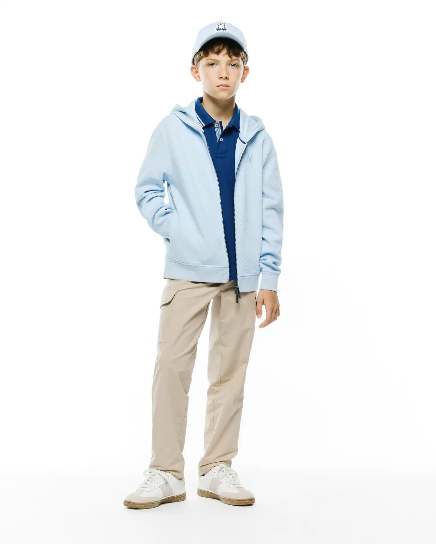 Kids Marc Textured Full Zip Hoodie in light blue, with textured fabric and an embroidered Psycho Bunny chest logo. Layered over a navy pique polo, beige cargo pants, and a light blue Psycho Bunny cap.