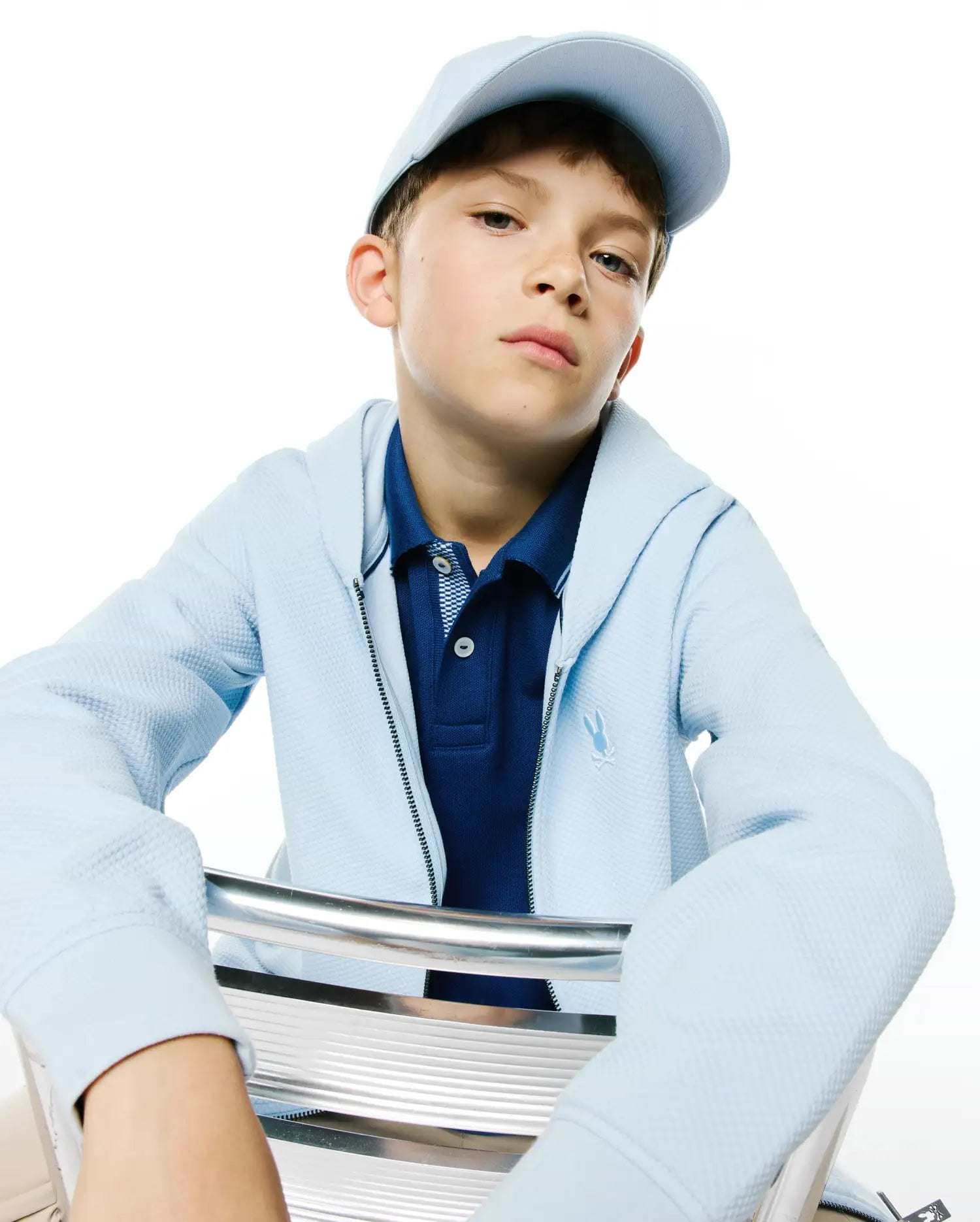 Kids Marc Textured Full Zip Hoodie in light blue, featuring a textured fabric and an embroidered Psycho Bunny skull logo on the left chest. Worn with a navy pique polo and a light blue cap.