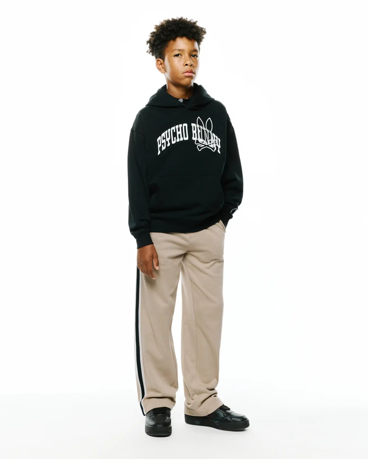 Black Kids Nathaniel Relaxed Hoodie, a fleece garment, featuring a large white Psycho Bunny skull logo chest graphic, paired with khaki woven relaxed fit pants with a black and white side stripe.