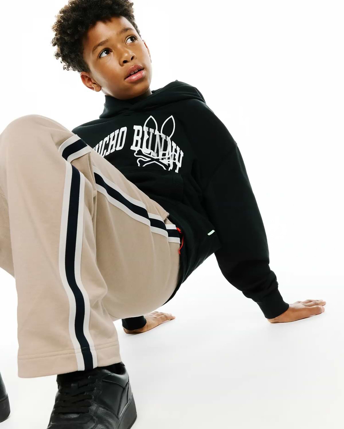 Kids Nathaniel Relaxed Hoodie in black french terry with a bold white Psycho Bunny skull logo print on the chest, complemented by relaxed beige track pants with black and white side stripes.