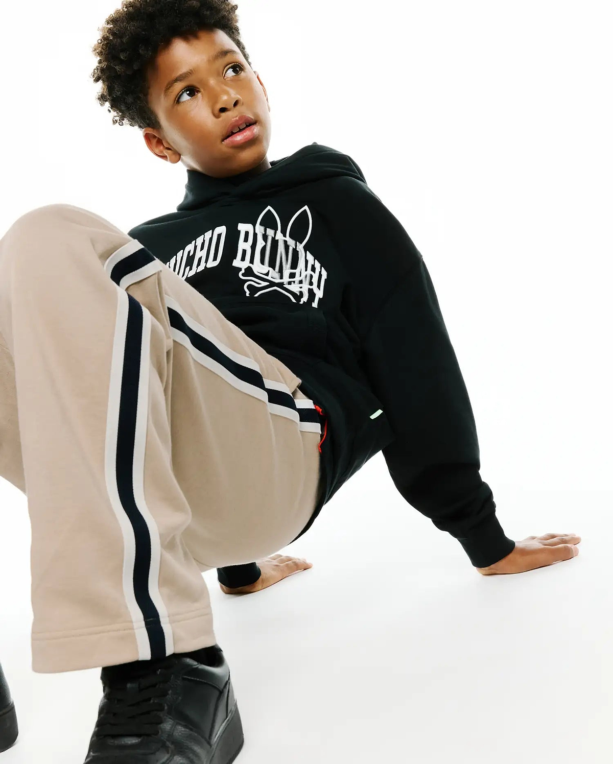 Kids Nathaniel Relaxed Hoodie in black french terry with a bold white Psycho Bunny skull logo print on the chest, complemented by relaxed beige track pants with black and white side stripes.