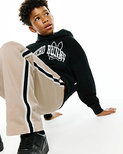 Kids Nathaniel Relaxed Hoodie in black french terry with a bold white Psycho Bunny skull logo print on the chest, complemented by relaxed beige track pants with black and white side stripes.