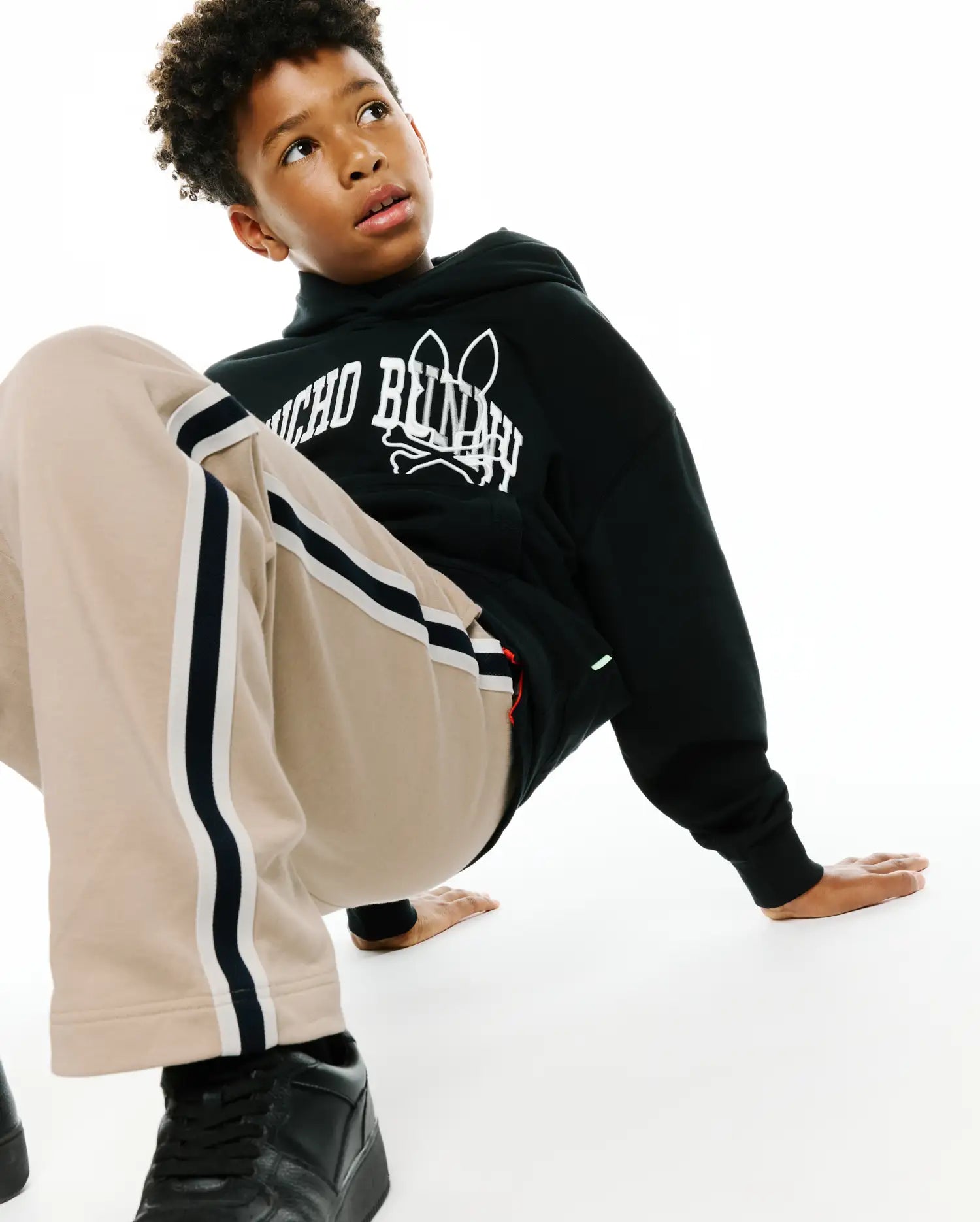 Kids Nathaniel Relaxed Hoodie in black french terry with a bold white Psycho Bunny skull logo print on the chest, complemented by relaxed beige track pants with black and white side stripes.