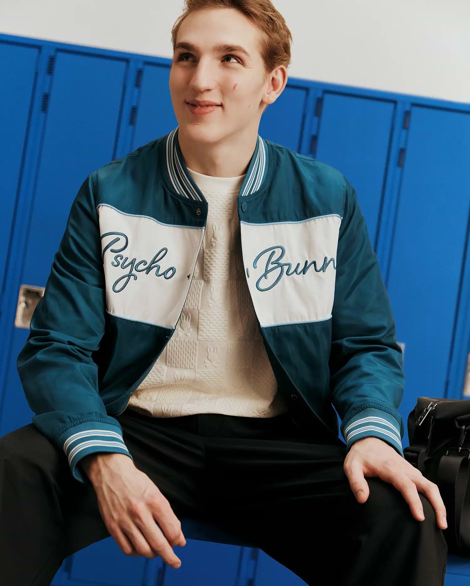 Mens Thomas Satin Bomber in deep teal with a white chest panel, featuring a large light blue embroidered Psycho Bunny script logo, and striped ribbed collar and cuffs for a classic fit.