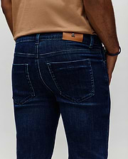 Mens Emerson Denim Pant in dark indigo blue denim, featuring a classic fit, subtle faded wash, light blue contrast stitching, and a small Psycho Bunny skull logo on a tan leather patch at the back waistband.