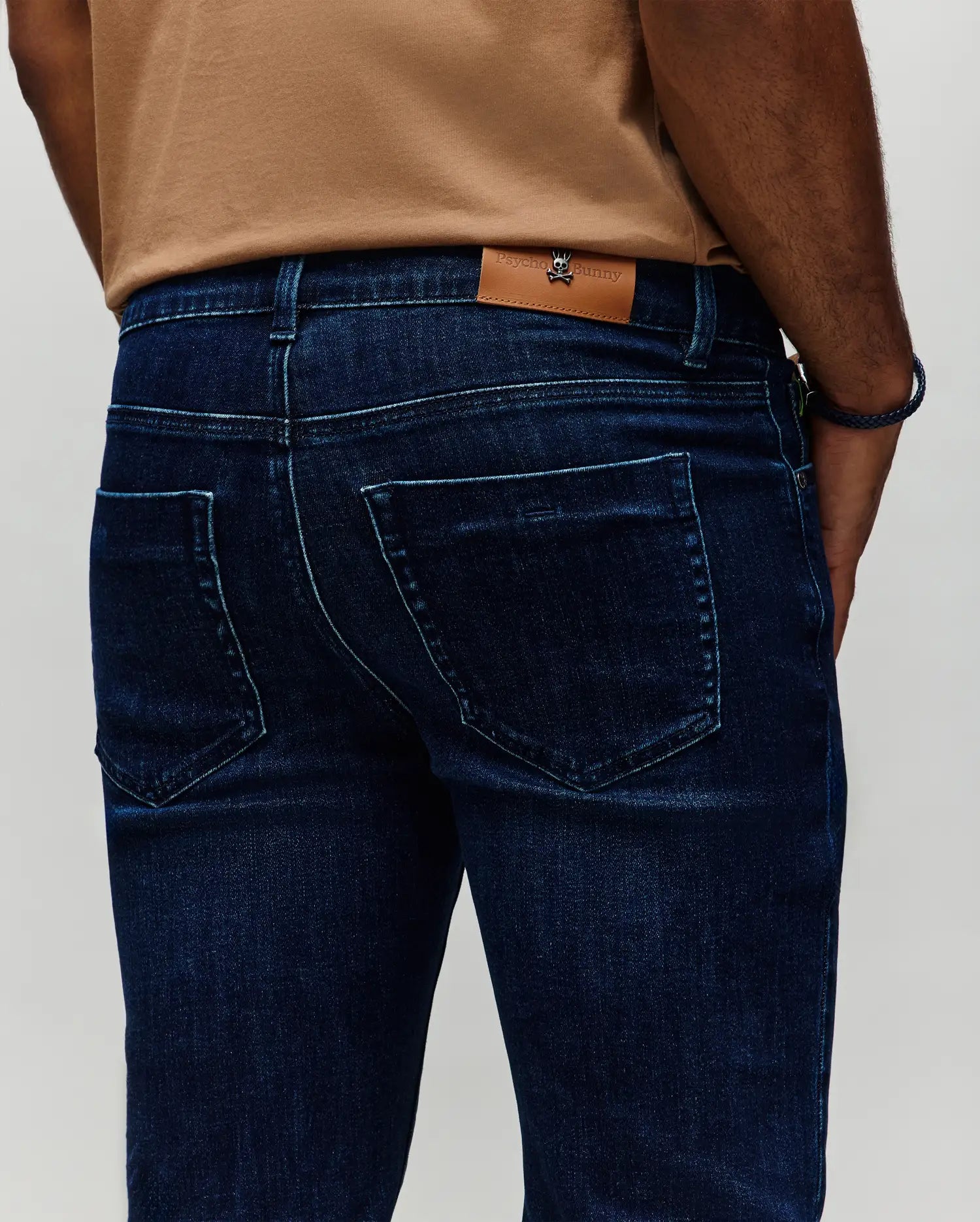 Mens Emerson Denim Pant in dark indigo blue denim, featuring a classic fit, subtle faded wash, light blue contrast stitching, and a small Psycho Bunny skull logo on a tan leather patch at the back waistband.