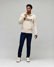 Cream knit hoodie with large brown embroidered Psycho Bunny skull logo and text on chest, kangaroo pocket, paired with dark blue slim Mens Emerson Denim Pant.