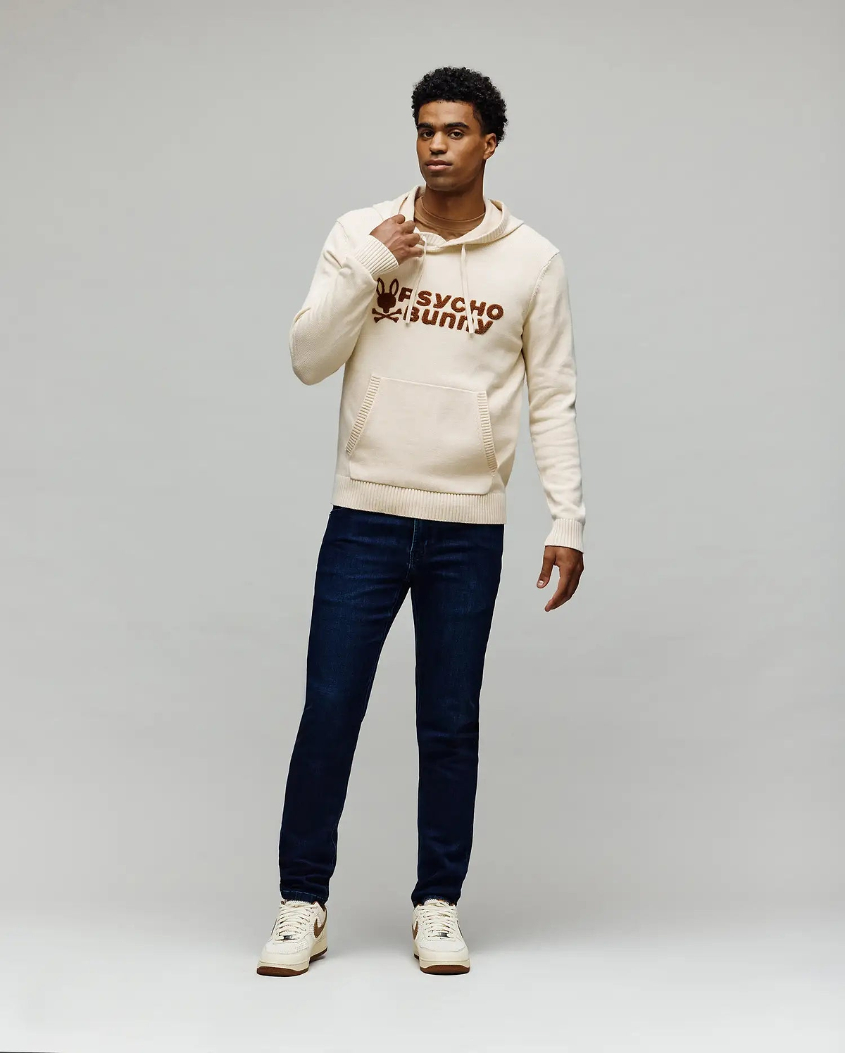 Cream knit hoodie with large brown embroidered Psycho Bunny skull logo and text on chest, kangaroo pocket, paired with dark blue slim Mens Emerson Denim Pant.