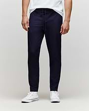 A person wears Psycho Bunny’s MENS BEN TECH JOGGER PULL ON (MB0600013) in navy, paired with a white T-shirt and white sneakers, standing against a plain light gray background. The photo is cropped from the chest down. (1)