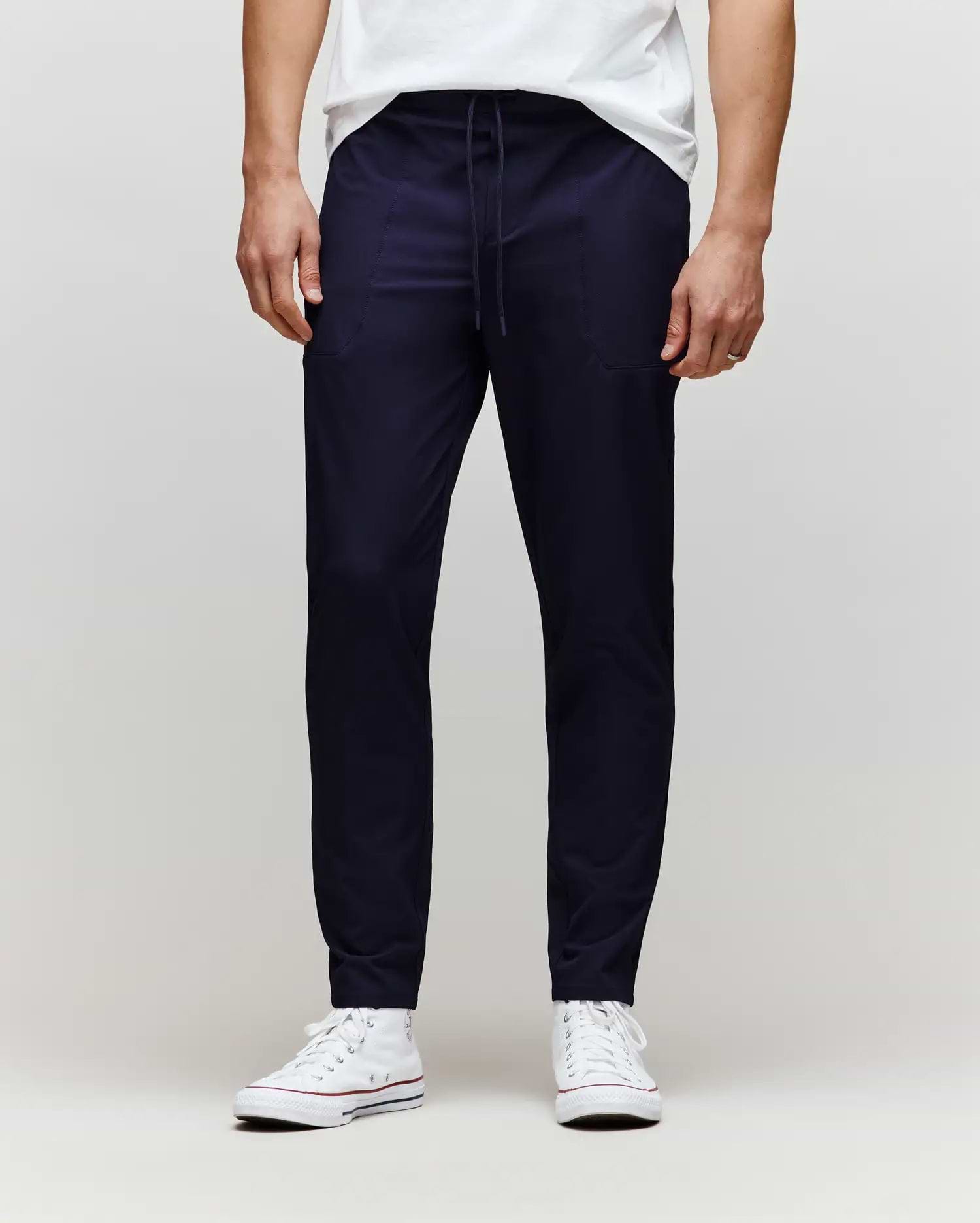 A person wears Psycho Bunny’s MENS BEN TECH JOGGER PULL ON (MB0600013) in navy, paired with a white T-shirt and white sneakers, standing against a plain light gray background. The photo is cropped from the chest down. (1)
