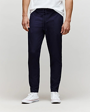 A person wears Psycho Bunny’s MENS BEN TECH JOGGER PULL ON (MB0600013) in navy, paired with a white T-shirt and white sneakers, standing against a plain light gray background. The photo is cropped from the chest down. (1)