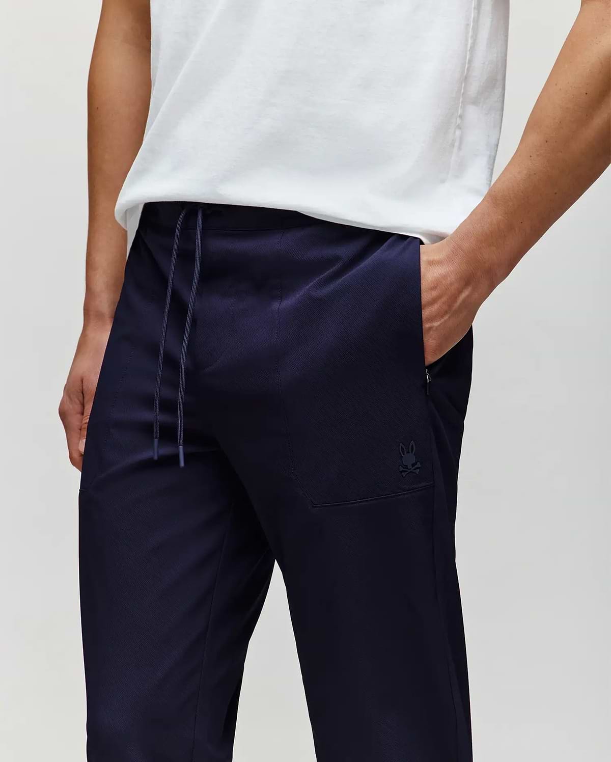 A person models Psycho Bunny’s MENS BEN TECH JOGGER PULL ON - MB0600013 in dark navy polyester spandex, shown from shoulders to mid-thigh with one hand in their pocket against a plain background. (2)