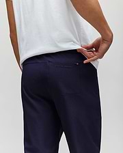 A man in a white shirt and PSYCHO BUNNY MENS BEN TECH JOGGER PULL ON pants (MB0600013) made from woven fabric stands with his back to the camera, pulling his shirt to show the "Psycho Bunny" logo above the back pocket. (3)