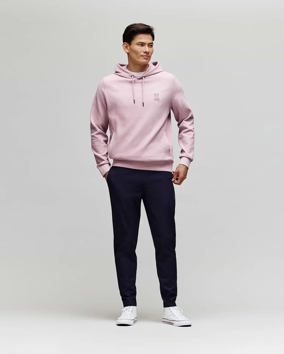 A man stands against a plain light background, wearing Psycho Bunny's MENS BEN TECH JOGGER PULL ON - MB0600013 in dark navy, paired with a light pink hoodie and white sneakers. He smiles slightly, hands in pockets, looking to the side. (4)