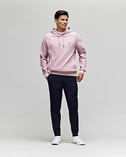 A man stands against a plain light background, wearing Psycho Bunny's MENS BEN TECH JOGGER PULL ON - MB0600013 in dark navy, paired with a light pink hoodie and white sneakers. He smiles slightly, hands in pockets, looking to the side. (4)