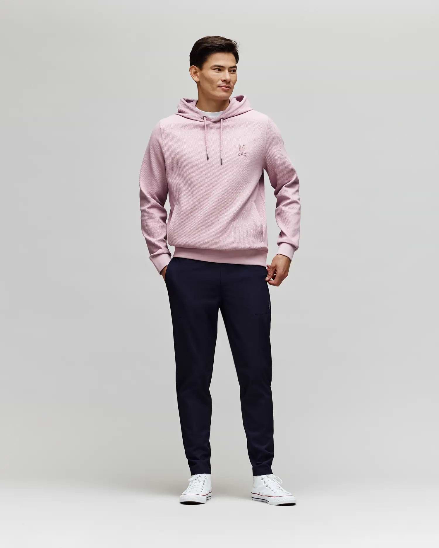 A man stands against a plain light background, wearing Psycho Bunny's MENS BEN TECH JOGGER PULL ON - MB0600013 in dark navy, paired with a light pink hoodie and white sneakers. He smiles slightly, hands in pockets, looking to the side. (4)