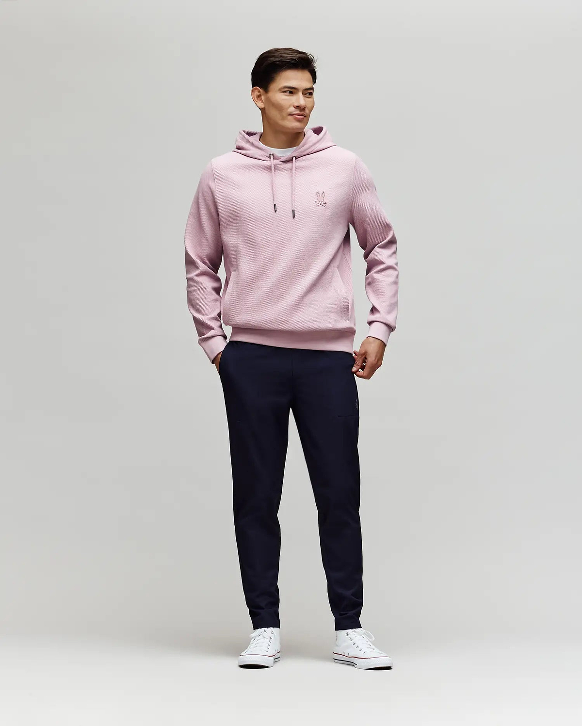 A man stands against a plain light background, wearing Psycho Bunny's MENS BEN TECH JOGGER PULL ON - MB0600013 in dark navy, paired with a light pink hoodie and white sneakers. He smiles slightly, hands in pockets, looking to the side. (4)